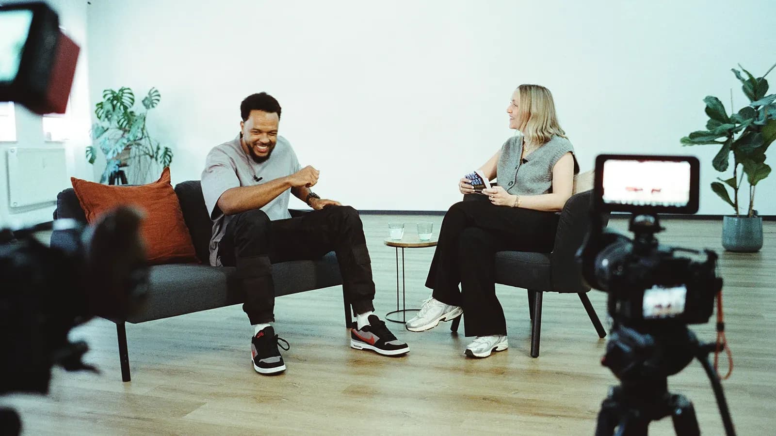 Zwei Personen im Interview auf einem Sofa, davor eine Kamera auf Stativ