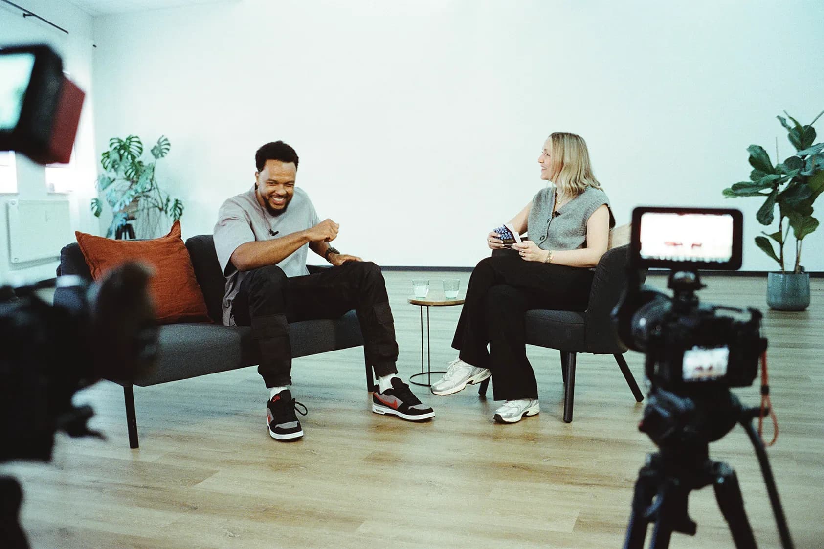 Zwei Personen im Interview auf einem Sofa, davor eine Kamera auf Stativ
