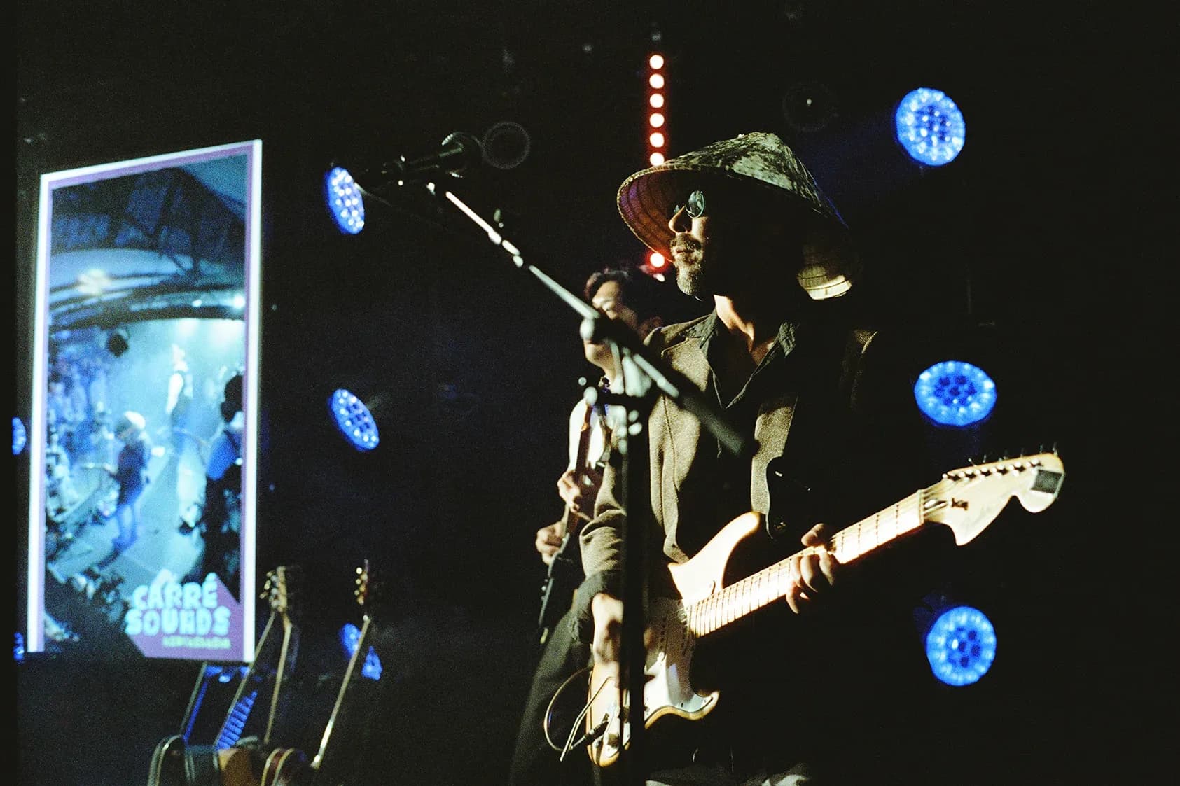 Gitarrist mit Hut im Gegenlicht, daneben die LED-Wand mit dem Carré-Sounds-Motiv